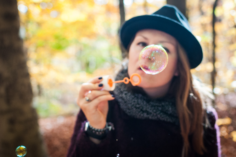 Raphaela in a dark sweater and hat blowing bubbles in an autumn forest, surrounded by golden foliage and soft sunlight, evoking playful healing and self-discovery
