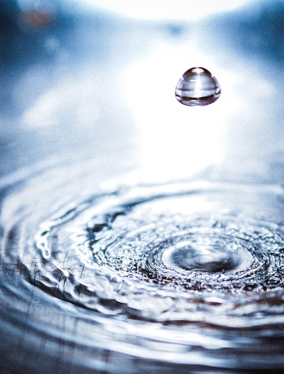 Single water droplet suspended above a calm surface with concentric ripples spreading outward, evoking reflection, cause and effect, and mindful transformation