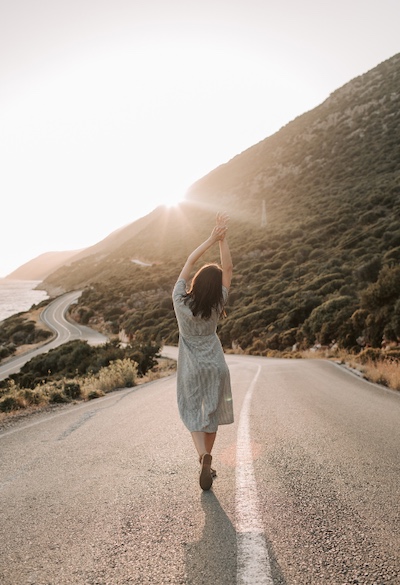 Person walking with arms raised on a winding road through a scenic, sunlit landscape, evoking emotional release, personal freedom, and reflective movement