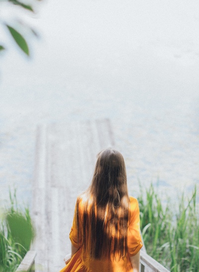 Person in an orange shirt heading towards the end of a dock, gazing over a misty lake surrounded by foliage, evoking reflection, solitude, and the threshold of change