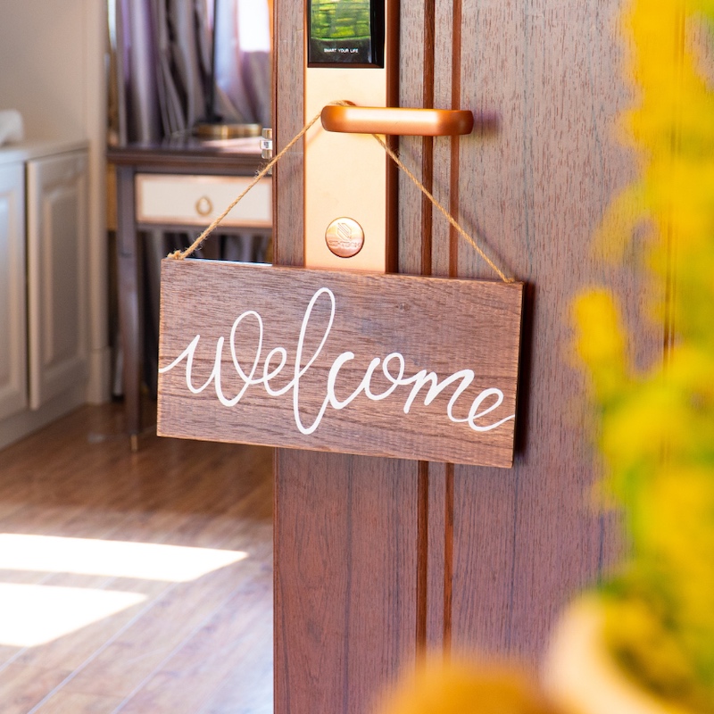 Wooden "welcome" sign hanging from a golden door handle, with a softly lit room visible in the background