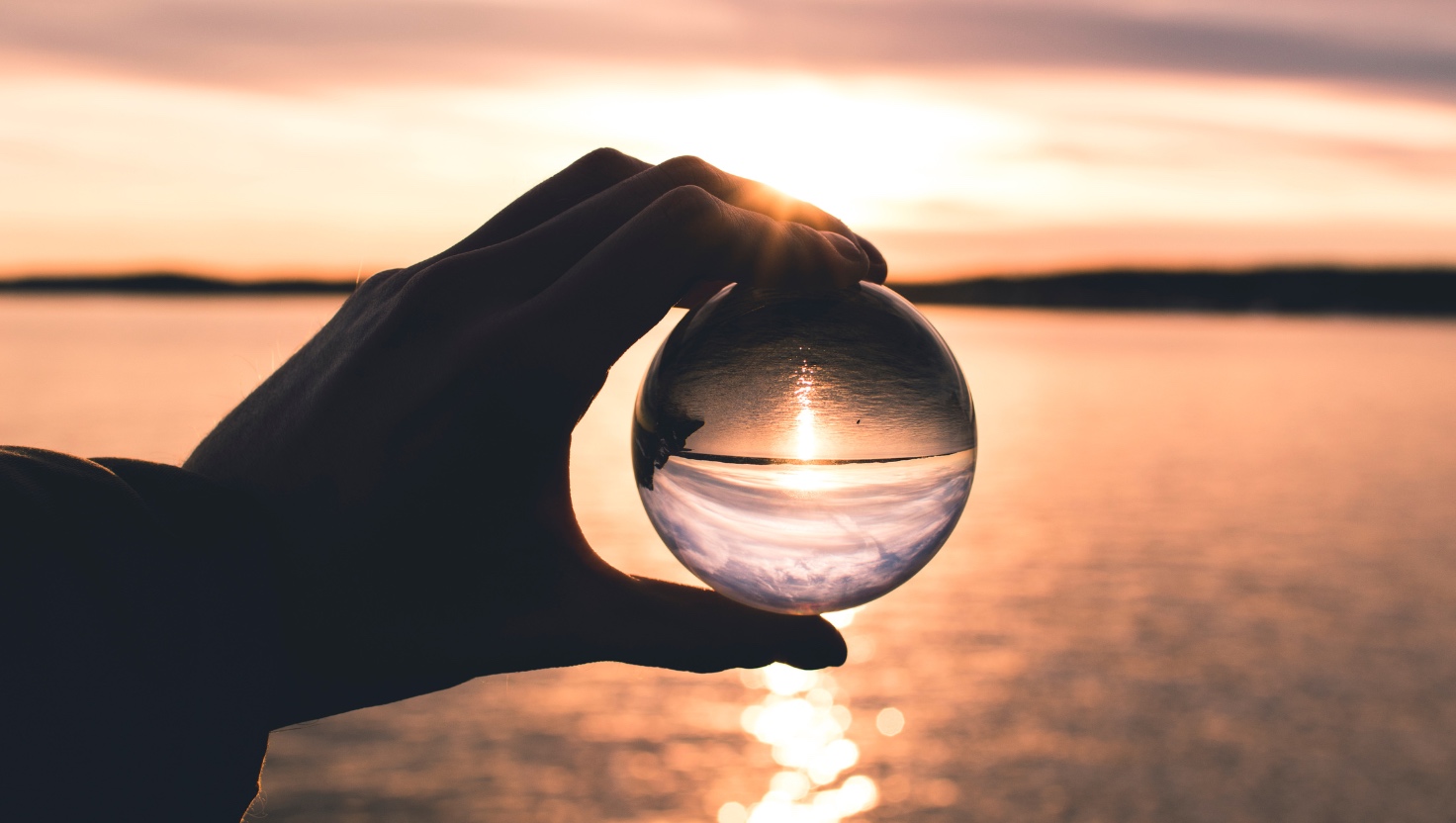 Silhouetted hand holding a glass sphere at sunset over water, with the inverted scene inside the sphere symbolizing reflection and new perspective.